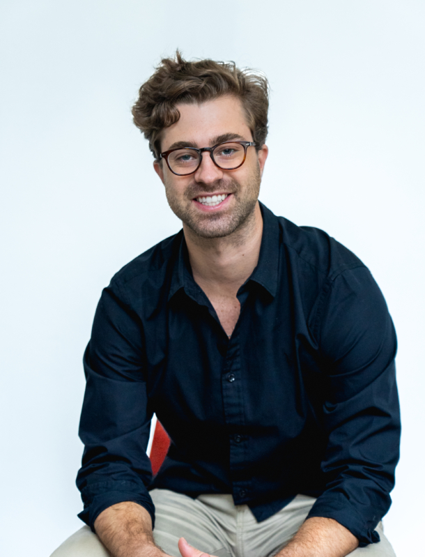 Alex sitting in front of a white background, smiling at the camera and wearing a black shirt with khaki pants.